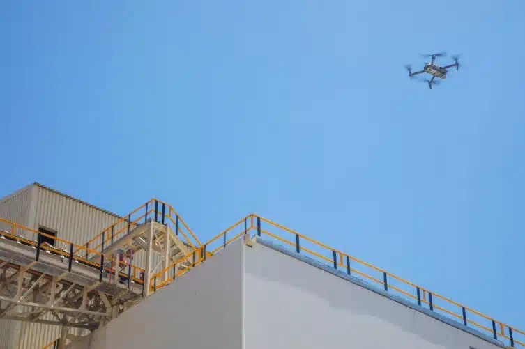 Drone lentää suuren teollisuusrakennuksen yllä, kirkasta sinistä taivasta vasten, esitellen modernia teknologiaa rakennusympäristössä, suorittaen ilmatarkastusta.